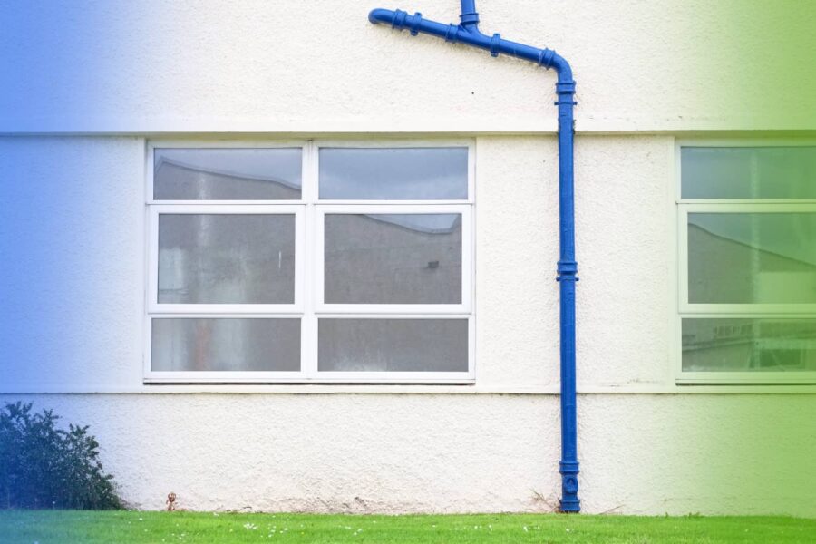A bright blue vertical pipe runs down the textured, off-white exterior wall of a building between two sets of windows.