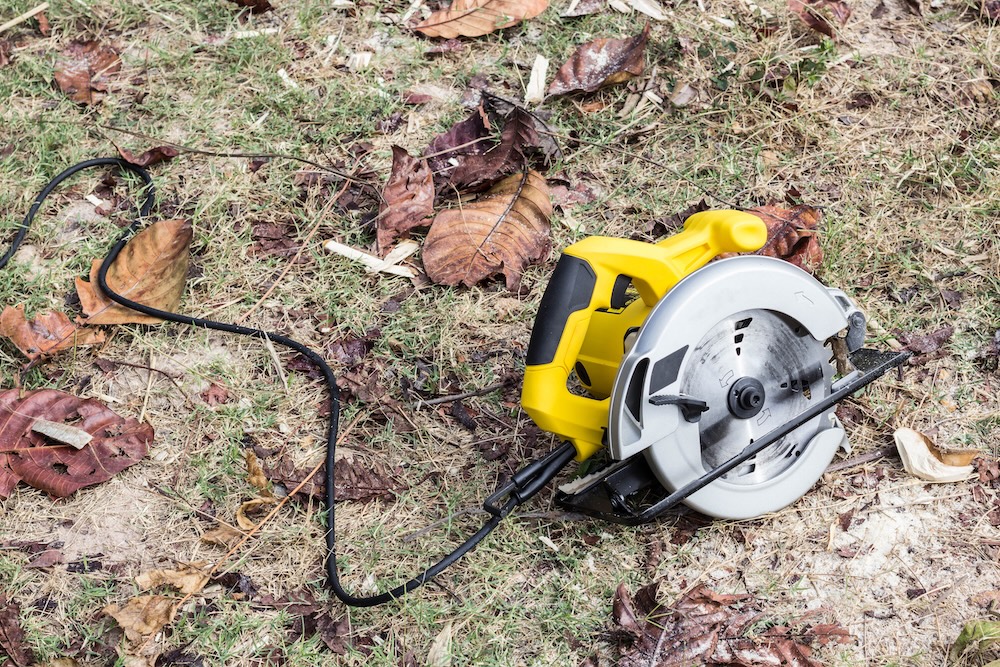 A yellow handheld circular saw resting on dry grass and fallen leaves with its power cord extended.