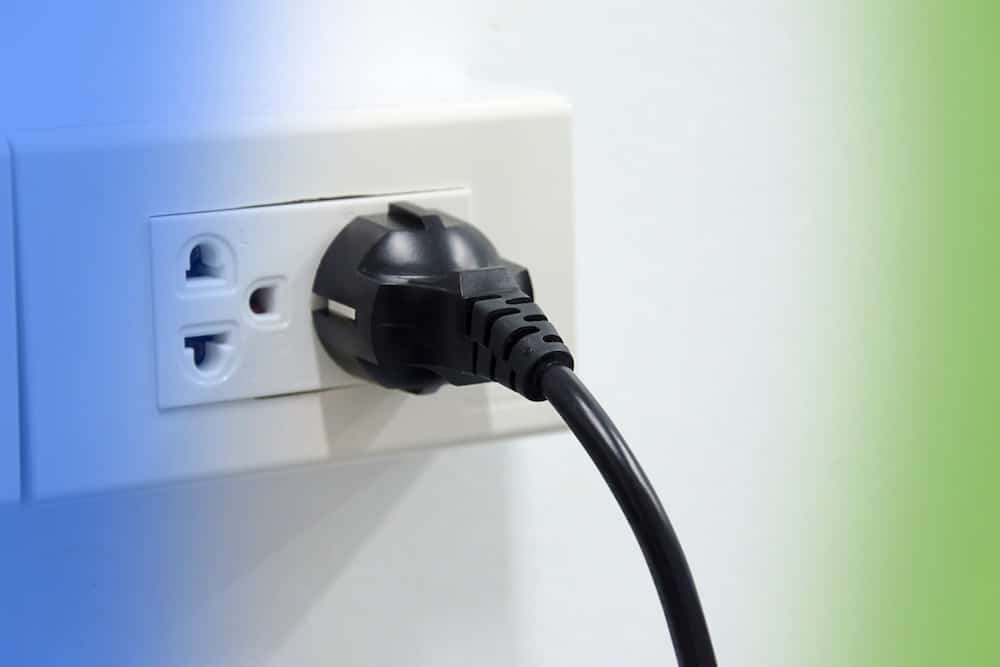 A black power plug with an attached cable is inserted into a white electrical wall outlet.
