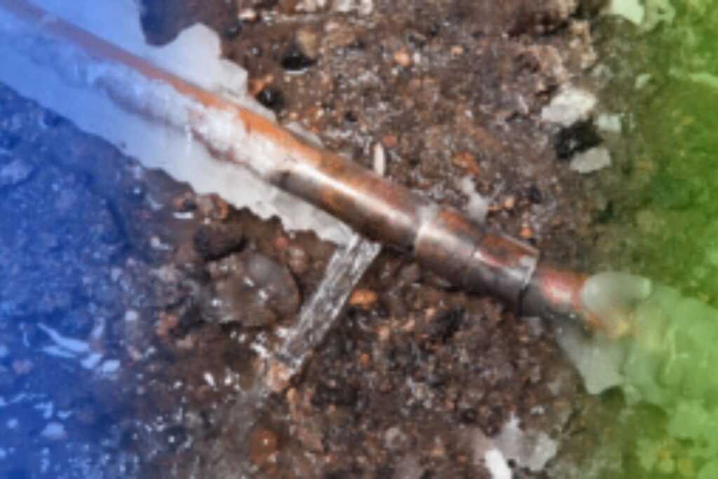Frozen copper water pipe encrusted in thick ice with a crack actively spraying a jet of water onto the ground.